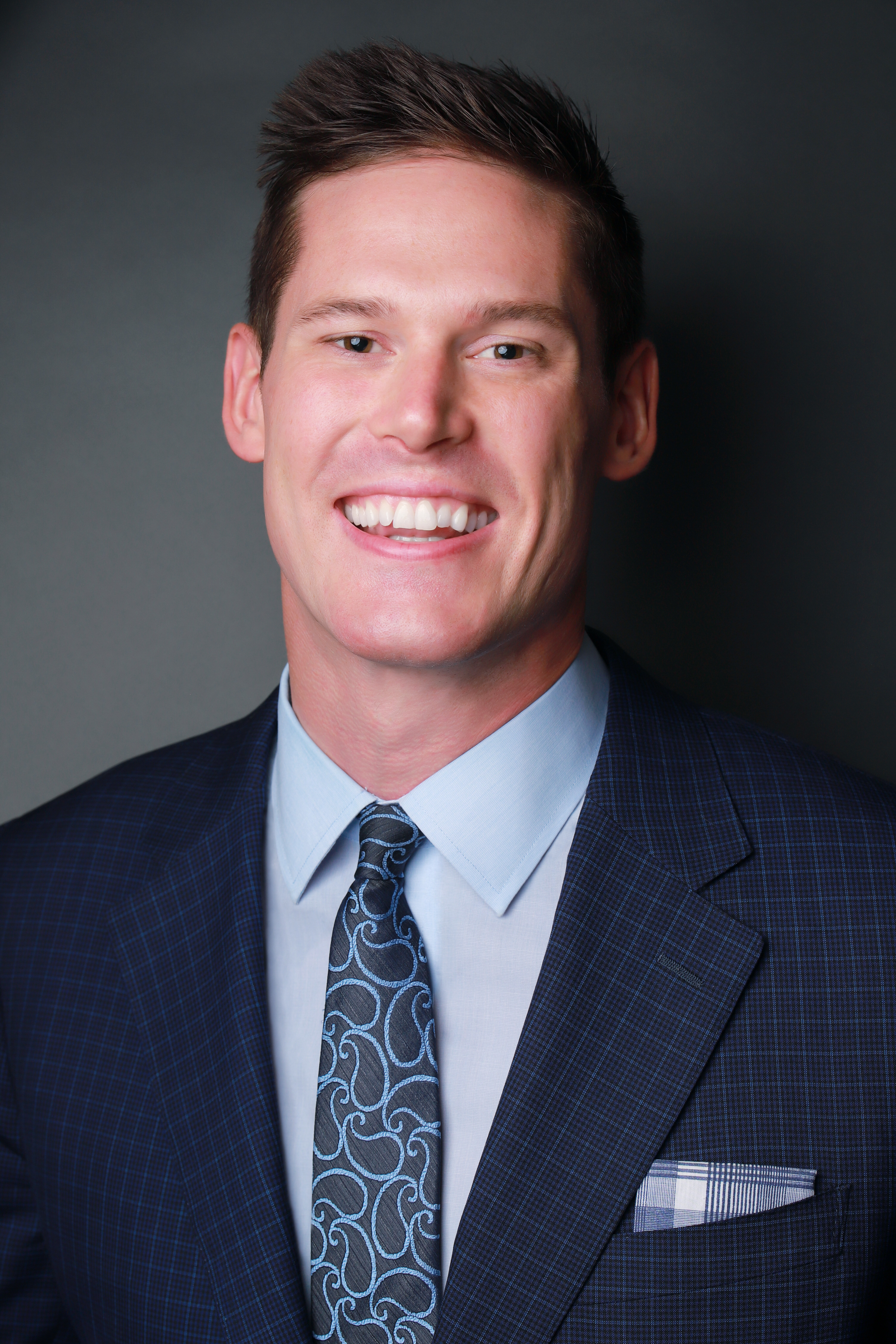 Young man smiling calmly in a dark blue suit and tie. His eyes look kind, competent, and trustworthy.